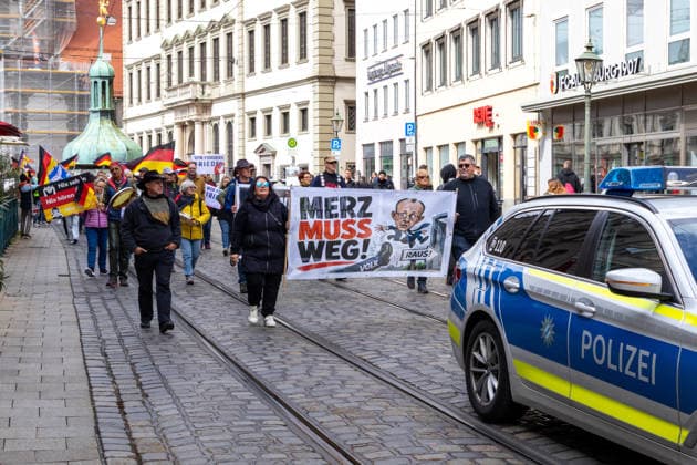 Demonstration gegen die aktuelle Bundesregierung unter Bundeskanzler Friedrich Merz in Augsburg. Unter dem Motto: Merz muss weg! Teilnehmer mit Banner mit Aufschrift: MERZ MUSS WEG! Mit Illustration von einem Fuß auf dem VOLK steht und einer Illustration von Bundeskanzler Friedrich Merz (CDU) der durch eine Tür fliegt mit einem Schild m