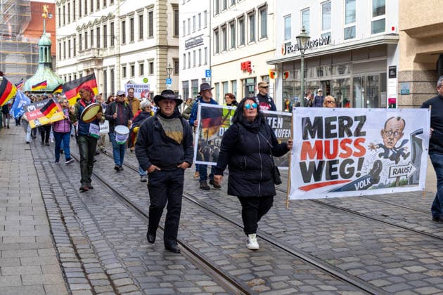 Demonstration gegen die aktuelle Bundesregierung unter Bundeskanzler Friedrich Merz in Augsburg. Unter dem Motto: Merz muss weg! Teilnehmer mit Banner mit Aufschrift: MERZ MUSS WEG! Mit Illustration von einem Fuß auf dem VOLK steht und einer Illustration von Bundeskanzler Friedrich Merz (CDU) der durch eine Tür fliegt mit einem Schild m