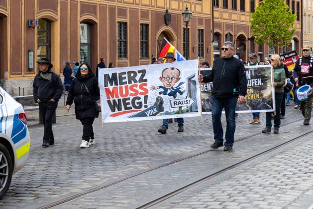 Demonstration gegen die aktuelle Bundesregierung unter Bundeskanzler Friedrich Merz in Augsburg. Unter dem Motto: Merz muss weg! Teilnehmer mit Banner mit Aufschrift: MERZ MUSS WEG! Mit Illustration von einem Fuß auf dem VOLK steht und einer Illustration von Bundeskanzler Friedrich Merz (CDU) der durch eine Tür fliegt mit einem Schild m