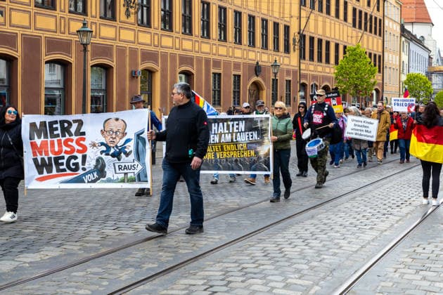 Demonstration gegen die aktuelle Bundesregierung unter Bundeskanzler Friedrich Merz in Augsburg. Unter dem Motto: Merz muss weg! Teilnehmer mit Banner mit Aufschrift: MERZ MUSS WEG! Mit Illustration von einem Fuß auf dem VOLK steht und einer Illustration von Bundeskanzler Friedrich Merz (CDU) der durch eine Tür fliegt mit einem Schild m