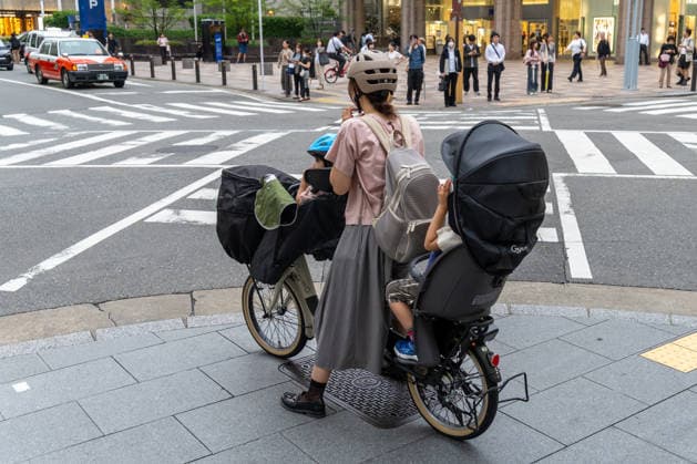 Eine Mutter transportiert in Japan Kinder mit einem Lastenrad sicher durch den urbanen Straßenverkehr. Das Bild steht für nachhaltige Mobilität, Familienalltag, Fahrradkultur und moderne Stadtentwicklung.