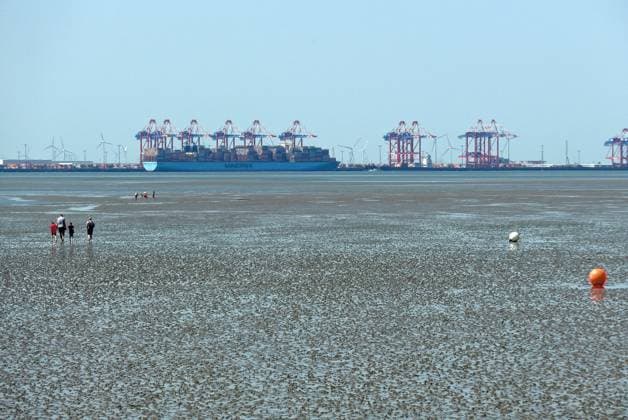 Blick vom Friesenstrand im Nordseebad Tossens, einem Ortsteil der Gemeinde Butjadingen im Landkreis Wesermarsch (Niedersachsen), auf die Nordseebucht Jadebusen mit trockenfallendem Watt. Im Hintergrund der Tiefwasserhafen Jade-Weser-Port (JWP) Wilhelmshaven. Das Wattenmeer gehört zum UNESCO-Weltnaturerbe.