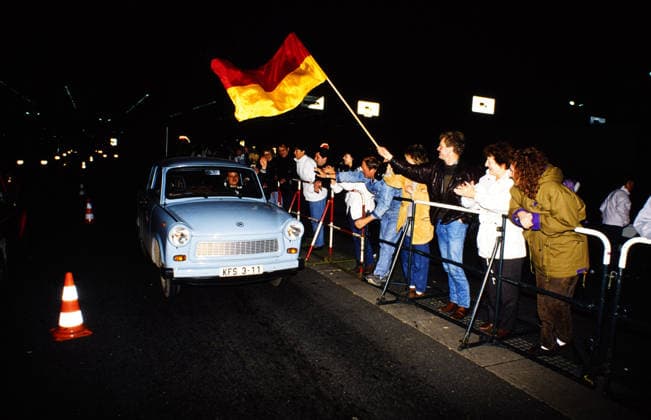 Ein hellblauer Trabant fährt nachts über die geöffnete Grenze, jubelnde Menschen mit einer Deutschlandfahne stehen am Straßenrand und begrüßen die Insassen, Deutschland, 1989. Berlin Berlin Deutschland Copyright: xUnitedxArchivesx xSvenxSimonxArchive SVS_94060000002