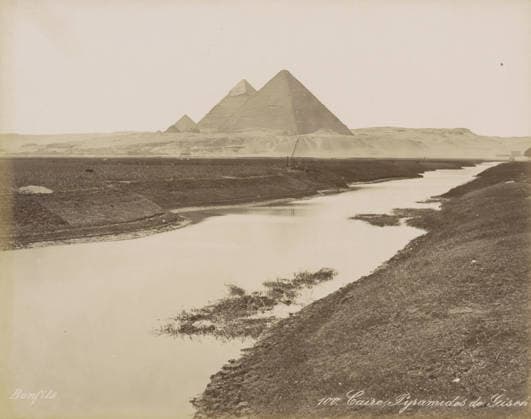 RECORD DATE NOT STATED Caire, Pyramids of Giza, View of the Pyramids of Giza near Cairo, photograph, Part of Travel Album with 80 Photos of Egypt., negative number: 100 , photographer: Maison Bonfils, Giza, c. 1870 - c. 1898, North Africa, pasteboard, paper, albumen print, height 220 mm x width 282 mm, pyramid (historical grave form), Ottoman Empire, Cairo (city), A wide sepia-toned landscape showing a calm winding river in the foreground with grassy and muddy banks, a low flat plain beyond the river, and three large pyramidal stone structures on a distant rise against a pale sky, with sparse vegetation and a small boat or wooden structure near the far bank.  Copyright Penta Springs LCK260531_03313