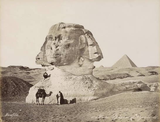 RECORD DATE NOT STATED Sphinx and pyramids of Giza, Caire, Sphynx et Pyramide, photograph, Part of Travel album with 80 photos of Egypt., negative number: 111 , photographer: Maison Bonfils, Gizeh, c. 1870 - c. 1898, North Africa, Giza, pasteboard, paper, albumen print, height 220 mm x width 282 mm, pyramid (historical grave form), piece of sculpture, reproduction of a piece of sculpture (sculpture), A large weathered stone sculpture of a human head and neck dominates the foreground, showing layered textures and erosion lines on its surface. A person sits on the stone near the sculpture™s base, and another person stands in front of the base beside a camel, which has a saddle and some coverings. A small figure is positioned on a ledge on the sculpture™s neck.  Copyright Penta Springs LCK260531_03315