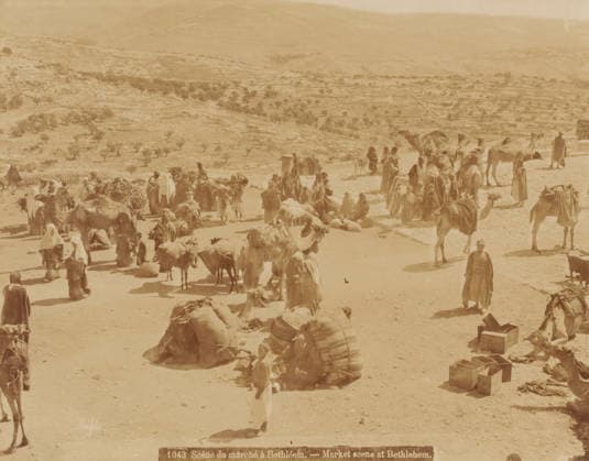 RECORD DATE NOT STATED Market scene in Bethlehem, Jerusalem, Palestine, photograph, Palestine., inscription: Bonfils. , inscription: 1043 Scene du marchà à Bethlà em. - Market scene at Bethlehem , photographer: Maison Bonfils, c. 1850 - c. 1900, paper, albumen print, height 217 mm x width 277 mm, height 351 mm x width 397 mm, A sunlit open area with many people wearing long robes and head coverings gathered among several camels and pack animals, some animals standing and some lying on the ground, bundles and packed saddles placed nearby, groups of people standing, walking, or sitting in small clusters, a few wooden crates and boxes arranged on the ground, a distant hilly landscape with scattered trees and terraces visible in the background, and a dusty groun Copyright Penta Springs LCK260531_17577