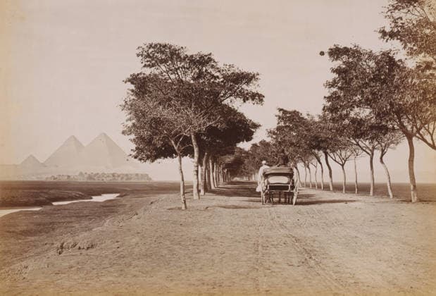 RECORD DATE NOT STATED Pyramid of Cheops, Grande pyramide de ChÃ ops, photograph, number: 113 , photographer: FÃ lix Bonfils, Giza, 1867 - 1885, North Africa, pasteboard, paper, albumen print, height 200 mm x width 279 mm, height 396 mm x width 290 mm, pyramid (historical grave form), A long straight dirt road lined with two rows of tall leafy trees forming a shaded avenue, a horse-drawn carriage with a covered back facing away from the viewer and two figures seated on and beside it, a narrow winding water channel running parallel to the road on the left, a cluster of low buildings beyond the channel, and three large pyramidal stone structures rising in the distant hazy background.  Copyright Penta Springs LCK260531_18458