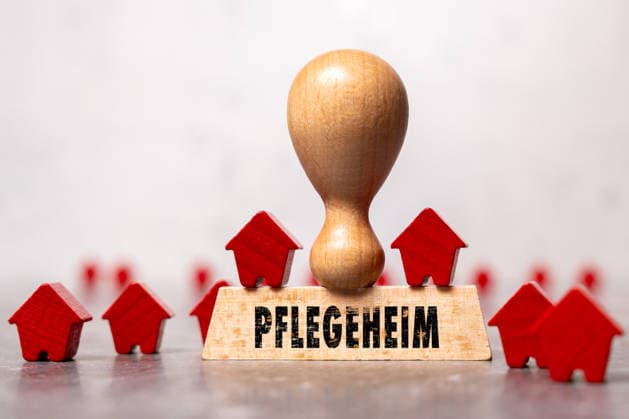 FOTOMONTAGE, Ein Holzstempel mit der Aufschrift Pflegeheim zwischen kleinen Holzhäusern symbolisiert stationäre Pflege und Seniorenwohnen. Das Motiv steht für Pflegeplätze, Betreuung, Demografiewandel und Pflegepolitik.
