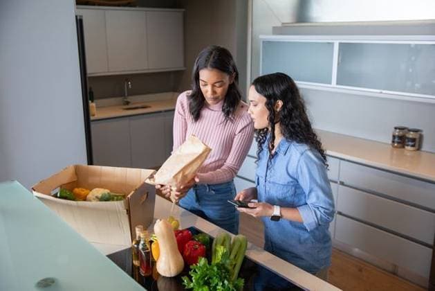 Diverse female friends unpacking cardboard box of veggies, checking smartphone at kitchen island, model released, property released, 09.04.2025 06:00:00, 6694377 Copyright: xIDS-250919_1xcreatorid:x0x 6694377