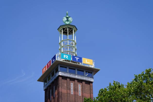 Detailaufnahme des Messeturms in Köln mit dem bunten RTL-Senderlogo und der markanten Turmspitze vor blauem Sommerhimmel mit grünen Blättern