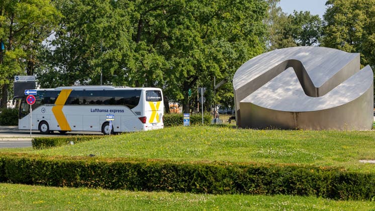 Flughafen Nürnberg, 04.08.2026 Ein Reisebus der Lufthansa Express fährt an einer markanten Metalllogo auf einem Kreisverkehr am Eingang des Flughafengeländes vorbei. Im Hintergrund ist ein Hinweisschild zum Flughafen zu erkennen. Nürnberg Flughafen Bayern Deutschland