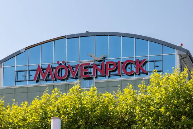 Flughafen Nürnberg, 04.08.2026 Gebäude eines Mövenpick Hotels mit markantem Logo direkt gegenüber dem Flughafenterminal. Die geschwungene Glasfassade spiegelt den blauen Himmel wider. Nürnberg Flughafen Bayern Deutschland