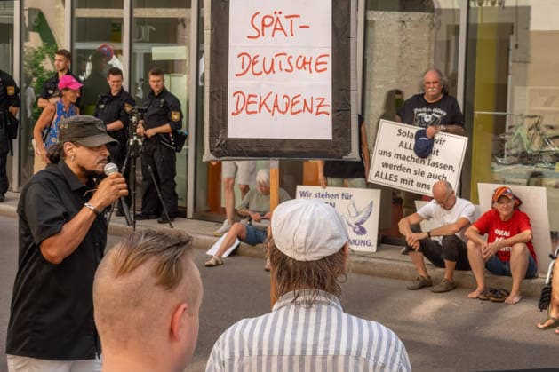 AfD-naher Protest gegen Dragqueen-Lesung in der Stadtbücherei Augsburg, ca. 30-40 Personen protestieren gegen Drag Queen Vicky Voyages Kinderbuch-Lesung in der Stadtbücherei, Aufruf: Jugendverein Aljuza, Augsburg, 5. August 2026 Deutschland, Augsburg, 5. August 2026, AfD-naher Protest gegen Dragqueen-Lesung in der Stadtbücherei Augsburg, Schild: Spät-deutsche Dekadenz, ca. 30-40 Personen protestieren gegen Drag Queen Vicky Voyage Kinderbuch-Lesung in der Stadtbücherei Augsburg, Ernst-Reuter-Platz, Mittwochnachmittag ab 15:30 Uhr, der AfD-nahe Jugendverein Aljuza demonstriert vor der Stadtbücherei gegen diesen Auftritt, Aljuza Abkürzung für Alternatives Jugendzentrum Augsburg, Gastredner Wiggins (phonetisch so verstanden, Vorname unk