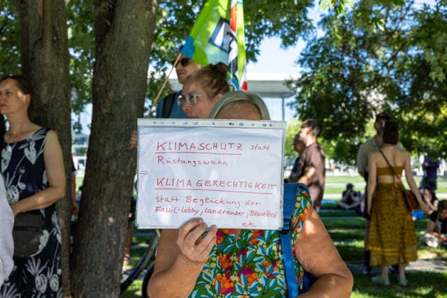 Fridays for Future Berlin - Hitzedemo. Deutschland, Berlin am 6. August 2026: Eine Frau hält ein kleines Schild hoch Klimaschutz statt Rüstungswahn. Klimagerechtigkeit statt Beglückung der Fossil-Lobby, Landräuber, Blackrock etc. Vor dem Kanzleramt protestieren Klimaaktivistinnen und Klimaaktivisten unter dem Motto Klimakrise eskaliert, Kanzler schweigt?!. Sie verlangen verstärkte Klimaschutzmaßnahmen, einen schnellen politischen Hitzeschutz sowie einen konsequenten Ausstieg aus fossilen Energien.
