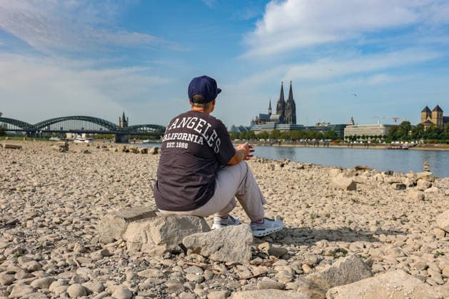 Sommer im Rheinland. Blick auf den Rhein bei Koeln (Köln) - Deutz. Der Rhein in Koeln (Köln) fuehrt (führt) Niedrigwasser aufgrund anhaltender Trockenheit. Im Hintergrund die Hohenzollernbruecke (Hohenzollernbrücke) (links) und der Koelner (Kölner) Dom (rechts). Im Vordergrund sitzt eine Frau auf einem Stein und schaut auf den Rhein. Sommer im Rheinland am 09.08.2026 in Koeln (Köln) Deutschland.