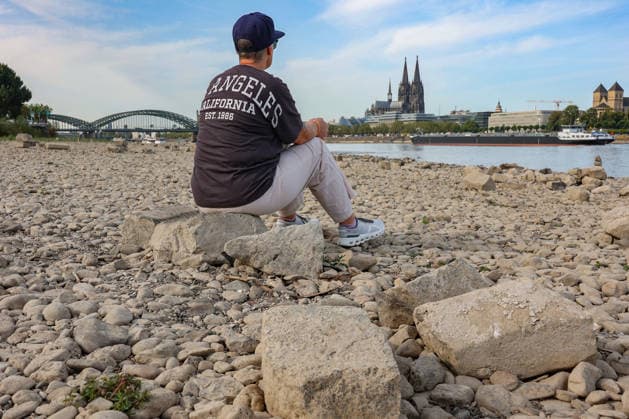 Sommer im Rheinland. Blick auf den Rhein bei Koeln (Köln) - Deutz. Der Rhein in Koeln (Köln) fuehrt (führt) Niedrigwasser aufgrund anhaltender Trockenheit. Im Hintergrund die Hohenzollernbruecke (Hohenzollernbrücke) (links) und der Koelner (Kölner) Dom (rechts). Im Vordergrund sitzt eine Frau auf einem Stein und schaut auf den Rhein. Sommer im Rheinland am 09.08.2026 in Koeln (Köln) Deutschland.
