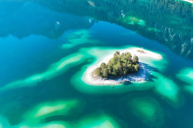 Wetterfeature, Morgenstille am Eibsee: Unterhalb der Zugspitze liegt der See bei Grainau nahe Garmisch-Partenkirchen in den frühen Stunden noch ruhig und nahezu unberührt. Doch an heißen Sommertagen ändert sich das Bild häufig schon ab Mittag. Dann strömen zahlreiche Ausflügler und Urlauber an den durch soziale Medien weltweit bekannt gewordenen Bergsee. Der zunehmende Massentourismus sorgt regelmäßig für einen regelrechten Besucheransturm. Unter dem hohen Verkehrsaufkommen, zugeparkten Straßen und dem großen Andrang leiden zunehmend auch die Anwohner in Grainau und rund um den Eibsee. (Aufnahme mit einer Drohne) Grainau Bayern Deutschland Grainau