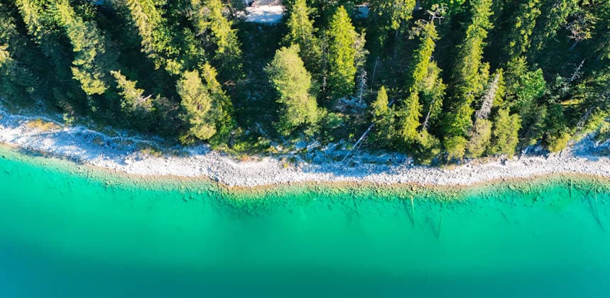 Wetterfeature, Morgenstille am Eibsee: Unterhalb der Zugspitze liegt der See bei Grainau nahe Garmisch-Partenkirchen in den frühen Stunden noch ruhig und nahezu unberührt. Doch an heißen Sommertagen ändert sich das Bild häufig schon ab Mittag. Dann strömen zahlreiche Ausflügler und Urlauber an den durch soziale Medien weltweit bekannt gewordenen Bergsee. Der zunehmende Massentourismus sorgt regelmäßig für einen regelrechten Besucheransturm. Unter dem hohen Verkehrsaufkommen, zugeparkten Straßen und dem großen Andrang leiden zunehmend auch die Anwohner in Grainau und rund um den Eibsee. (Aufnahme mit einer Drohne) Grainau Bayern Deutschland Grainau