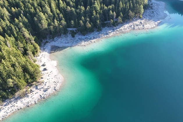 Wetterfeature, Morgenstille am Eibsee: Unterhalb der Zugspitze liegt der See bei Grainau nahe Garmisch-Partenkirchen in den frühen Stunden noch ruhig und nahezu unberührt. Doch an heißen Sommertagen ändert sich das Bild häufig schon ab Mittag. Dann strömen zahlreiche Ausflügler und Urlauber an den durch soziale Medien weltweit bekannt gewordenen Bergsee. Der zunehmende Massentourismus sorgt regelmäßig für einen regelrechten Besucheransturm. Unter dem hohen Verkehrsaufkommen, zugeparkten Straßen und dem großen Andrang leiden zunehmend auch die Anwohner in Grainau und rund um den Eibsee. (Aufnahme mit einer Drohne) Grainau Bayern Deutschland Grainau