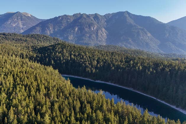 Wetterfeature, Morgenstille am Eibsee: Unterhalb der Zugspitze liegt der See bei Grainau nahe Garmisch-Partenkirchen in den frühen Stunden noch ruhig und nahezu unberührt. Doch an heißen Sommertagen ändert sich das Bild häufig schon ab Mittag. Dann strömen zahlreiche Ausflügler und Urlauber an den durch soziale Medien weltweit bekannt gewordenen Bergsee. Der zunehmende Massentourismus sorgt regelmäßig für einen regelrechten Besucheransturm. Unter dem hohen Verkehrsaufkommen, zugeparkten Straßen und dem großen Andrang leiden zunehmend auch die Anwohner in Grainau und rund um den Eibsee. (Aufnahme mit einer Drohne) Grainau Bayern Deutschland Grainau