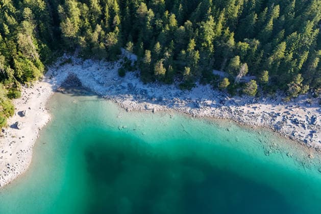 Wetterfeature, Morgenstille am Eibsee: Unterhalb der Zugspitze liegt der See bei Grainau nahe Garmisch-Partenkirchen in den frühen Stunden noch ruhig und nahezu unberührt. Doch an heißen Sommertagen ändert sich das Bild häufig schon ab Mittag. Dann strömen zahlreiche Ausflügler und Urlauber an den durch soziale Medien weltweit bekannt gewordenen Bergsee. Der zunehmende Massentourismus sorgt regelmäßig für einen regelrechten Besucheransturm. Unter dem hohen Verkehrsaufkommen, zugeparkten Straßen und dem großen Andrang leiden zunehmend auch die Anwohner in Grainau und rund um den Eibsee. (Aufnahme mit einer Drohne) Grainau Bayern Deutschland Grainau