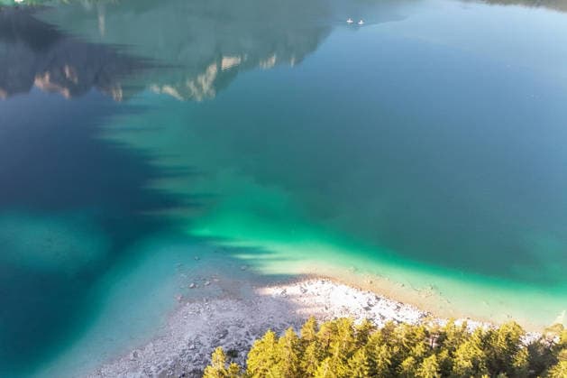 Wetterfeature, Morgenstille am Eibsee: Unterhalb der Zugspitze liegt der See bei Grainau nahe Garmisch-Partenkirchen in den frühen Stunden noch ruhig und nahezu unberührt. Doch an heißen Sommertagen ändert sich das Bild häufig schon ab Mittag. Dann strömen zahlreiche Ausflügler und Urlauber an den durch soziale Medien weltweit bekannt gewordenen Bergsee. Der zunehmende Massentourismus sorgt regelmäßig für einen regelrechten Besucheransturm. Unter dem hohen Verkehrsaufkommen, zugeparkten Straßen und dem großen Andrang leiden zunehmend auch die Anwohner in Grainau und rund um den Eibsee. (Aufnahme mit einer Drohne) Grainau Bayern Deutschland Grainau