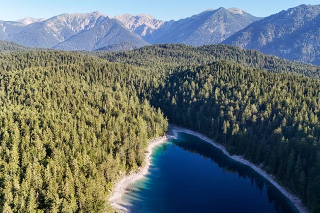Wetterfeature, Morgenstille am Eibsee: Unterhalb der Zugspitze liegt der See bei Grainau nahe Garmisch-Partenkirchen in den frühen Stunden noch ruhig und nahezu unberührt. Doch an heißen Sommertagen ändert sich das Bild häufig schon ab Mittag. Dann strömen zahlreiche Ausflügler und Urlauber an den durch soziale Medien weltweit bekannt gewordenen Bergsee. Der zunehmende Massentourismus sorgt regelmäßig für einen regelrechten Besucheransturm. Unter dem hohen Verkehrsaufkommen, zugeparkten Straßen und dem großen Andrang leiden zunehmend auch die Anwohner in Grainau und rund um den Eibsee. (Aufnahme mit einer Drohne) Grainau Bayern Deutschland Grainau