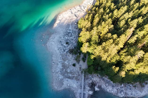 Wetterfeature, Morgenstille am Eibsee: Unterhalb der Zugspitze liegt der See bei Grainau nahe Garmisch-Partenkirchen in den frühen Stunden noch ruhig und nahezu unberührt. Doch an heißen Sommertagen ändert sich das Bild häufig schon ab Mittag. Dann strömen zahlreiche Ausflügler und Urlauber an den durch soziale Medien weltweit bekannt gewordenen Bergsee. Der zunehmende Massentourismus sorgt regelmäßig für einen regelrechten Besucheransturm. Unter dem hohen Verkehrsaufkommen, zugeparkten Straßen und dem großen Andrang leiden zunehmend auch die Anwohner in Grainau und rund um den Eibsee. (Aufnahme mit einer Drohne) Grainau Bayern Deutschland Grainau