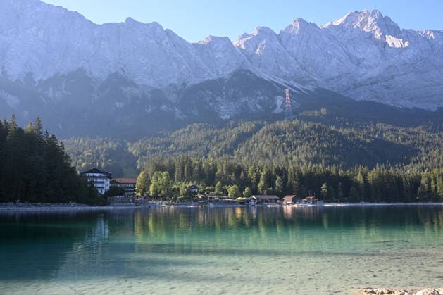 Wetterfeature, Morgenstille am Eibsee: Unterhalb der Zugspitze liegt der See bei Grainau nahe Garmisch-Partenkirchen in den frühen Stunden noch ruhig und nahezu unberührt. Doch an heißen Sommertagen ändert sich das Bild häufig schon ab Mittag. Dann strömen zahlreiche Ausflügler und Urlauber an den durch soziale Medien weltweit bekannt gewordenen Bergsee. Der zunehmende Massentourismus sorgt regelmäßig für einen regelrechten Besucheransturm. Unter dem hohen Verkehrsaufkommen, zugeparkten Straßen und dem großen Andrang leiden zunehmend auch die Anwohner in Grainau und rund um den Eibsee. Grainau Bayern Deutschland Grainau