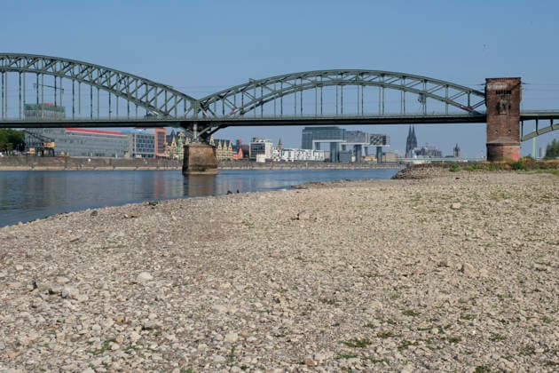 Eine bodennahe Aufnahme zeigt das weitläufige, freigelegte Kiesel- und Steinufer des Rheins bei extremem sommerlichem Niedrigwasser. Im unscharfen Hintergrund erstreckt sich die stählerne Fachwerk-Konstruktion der historischen Südbrücke mit ihren markanten Wehrtürmen aus Backstein. Dahinter zeichnet sich die Kölner Skyline mit den modernen Kranhäusern im Rheinauhafen und den entfernten Spitzen des Kölner Doms unter einem klaren, blauen Himmel ab.