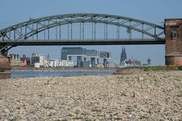 Eine bodennahe Aufnahme zeigt das weitläufige, freigelegte Kiesel- und Steinufer des Rheins bei extremem sommerlichem Niedrigwasser. Im unscharfen Hintergrund erstreckt sich die stählerne Fachwerk-Konstruktion der historischen Südbrücke mit ihren markanten Wehrtürmen aus Backstein. Dahinter zeichnet sich die Kölner Skyline mit den modernen Kranhäusern im Rheinauhafen und den entfernten Spitzen des Kölner Doms unter einem klaren, blauen Himmel ab.