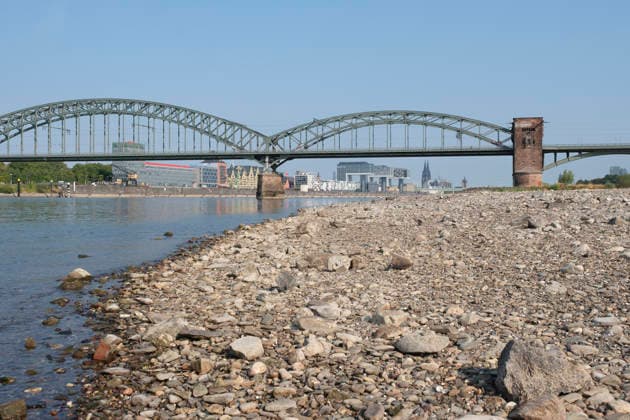 Eine weitwinklige Totalansicht zeigt den stark zurückgewichenen Flusslauf des Rheins mit einem weitläufig ausgetrockneten Kiesel- und Steinufer bei extremem sommerlichem Niedrigwasser. Die historische, stählerne Südbrücke überspannt den Fluss mit ihren charakteristischen Fachwerkbögen und dem markanten Wehrturm aus rotem Backstein. Im Hintergrund spiegelt sich das gegenüberliegende Rheinufer mit der Kölner Skyline, den Kranhäusern im Rheinauhafen und den entfernten Türmen des Kölner Doms unter einem wolkenlosen, blauen Himmel.