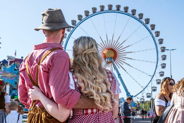 Oktoberfest 2026, Wiesn-Start bei strahlendem Sonnenschein und Temperaturen bis 24 Grad, bestes Wiesnwetter, Paar in Tracht vor dem Riesenrad, Eröffnungstag, 19. September 2026 Deutschland, München, 19. September 2026, Oktoberfest 2026, Wieesn-Start bei strahlendem Sonnenschein und Temperaturen bis 24 Grad, zwei Touristen aus Australien stilecht in Tracht vor dem Riesenrad, mittags um 12:30 kurz nach der Eröffnung, blauer Himmel, bestes Wiesnwetter, typisch bayerisch, Volksfest, Bayern,