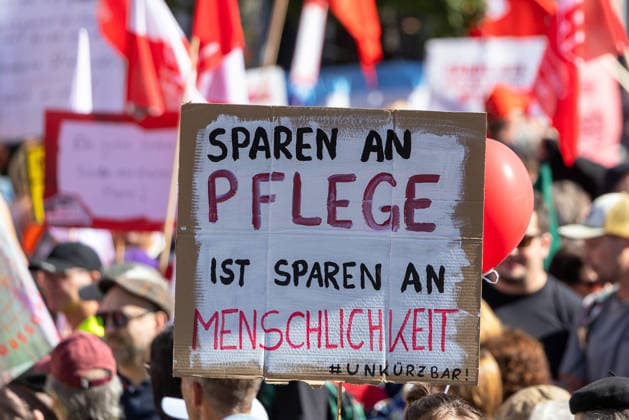 DGB Bayern - Hart verdient! Groß-Kundgebung, Nürnberg, 26.09.2026 Demonstranten halten bei einer Kundgebung ein Schild mit der Aufschrift Sparen an Pflege ist Sparen an Menschlichkeit und dem Hashtag Unkürzbar in die Höhe. Im Hintergrund sind zahlreiche weitere Teilnehmer mit roten Fahnen und Transparenten versammelt. Nürnberg Bayern Deutschland