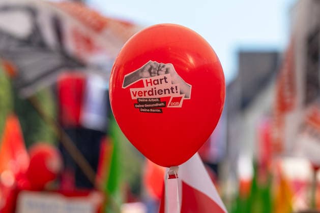 DGB Bayern - Hart verdient! Groß-Kundgebung, Nürnberg, 26.09.2026 Ein roter Luftballon mit der Aufschrift Hart verdient! Deine Arbeit. Deine Gesundheit. Deine Rente. und DGB-Logo ragt über eine Menschenmenge. Im unscharfen Hintergrund sind zahlreiche rote Fahnen einer gewerkschaftlichen Kundgebung zu erkennen. Nürnberg Bayern Deutschland