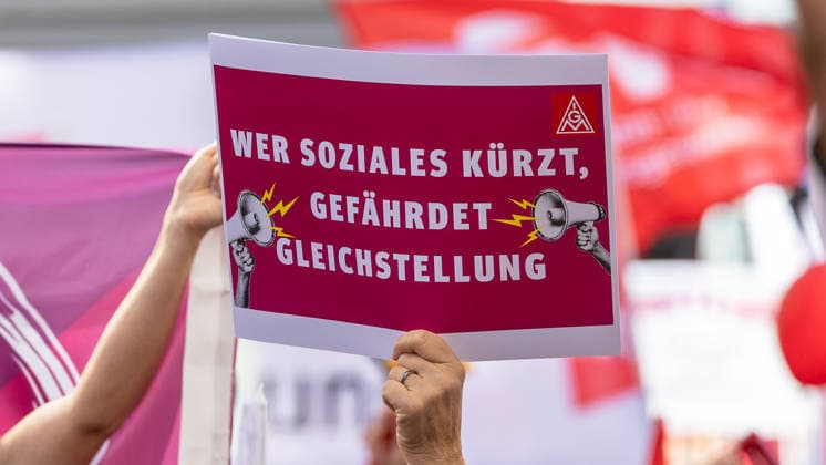 DGB Bayern - Hart verdient! Groß-Kundgebung, Nürnberg, 26.09.2026 Ein Demonstrant hält ein Schild der IG Metall mit der Aufschrift Wer Soziales kürzt, gefährdet Gleichstellung in die Höhe. Im Hintergrund sind weitere rote Fahnen und Transparente einer Kundgebung zu erkennen. Nürnberg Bayern Deutschland