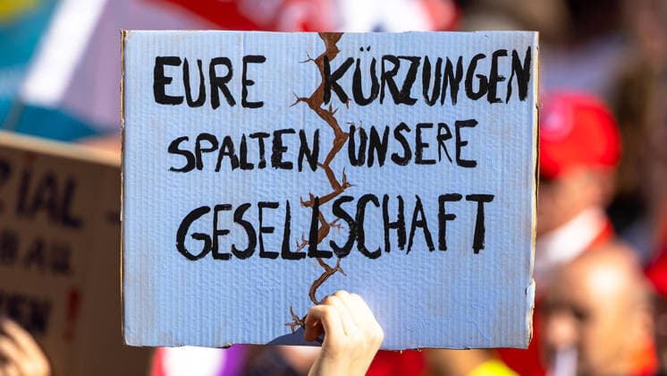 DGB Bayern - Hart verdient! Groß-Kundgebung, Nürnberg, 26.09.2026 Ein Demonstrant hält ein Pappschild mit der Aufschrift Eure Kürzungen spalten unsere Gesellschaft in die Höhe. Ein aufgemalter Riss zieht sich symbolisch durch die Mitte des Schildes, die Hand hält es entschlossen fest. Nürnberg Bayern Deutschland