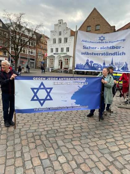 Flensburg, Schleswig-Holstein Flensburg gedenkt den Opfern des 7. Oktober 2023. Hier: Personen entfalten Transparente mit farblich weiß-blauen Hintergrund. LInks: Judenstern und Text Gegen jeden Antisemitismus, immer und überall! Für Respekt, Menschlichkeit und Zusammenhalt. Rechts: Jüdisches Leben in Flensburg. Sicher - sichtbar - selbstverständlich. Aufnahme vom 07.10.2026, Flensburg