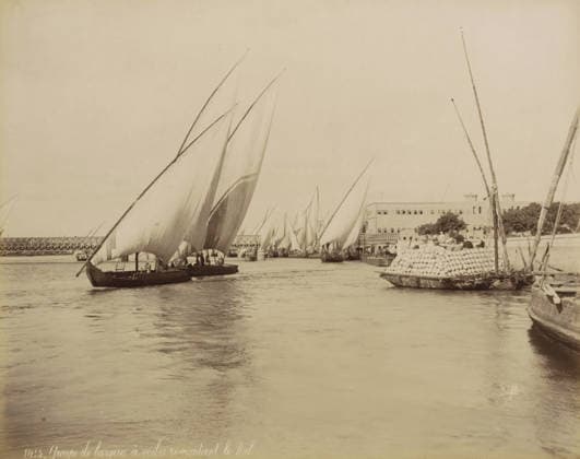 RECORD DATE NOT STATED Sailing boats with cargo on the Nile, Group of sailing boats sailing up the Nile, photograph, Part of Travel Album with 80 Photos of Egypt., negative number: 1415 , photographer: Maison Bonfils, Egypt, c. 1870 - c. 1898, North Africa, pasteboard, paper, albumen print, height 223 mm x width 280 mm, sailing-ship, sailing-boat, Nile, A wide water scene filled with multiple sailboats with large triangular sails, some sails overlapping and billowing, several small dark-hulled boats close to the camera and more farther back, a larger vessel at the right edge stacked high with rounded objects, a long low building with rows of windows along the shoreline, a metal bridge visible in the distance, vertical masts and poles rising from some boats, calm Copyright Penta Springs LCK260531_03318
