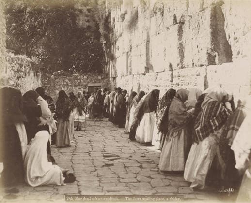 RECORD DATE NOT STATED Believes praying at the Wailing Wall in Jerusalem, Mur des Juifs un vendredi. - The Jews waiting place, a Friday., photograph, Part of Travel album with photos of Jerusalem and other biblical places., negative number: 245 , photographer: Maison Bonfils, Jerusalem, c. 1867 - c. 1895, pasteboard, paper, albumen print, height 0 mm x width 0 mm, prayer, praying, Jewish religion, Wailing Wall, A narrow stone-paved alley flanked on the right by a tall, light-colored stone wall and on the left by a lower rough stone wall and foliage, with a group of people standing or kneeling along the right wall and several others scattered along the left; many figures wear head coverings and layered garments with patterned shawls, some faces visible while othe Copyright Penta Springs LCK260531_03381