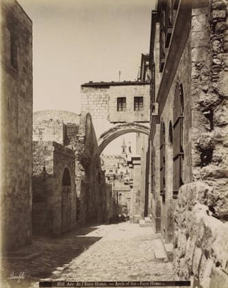 RECORD DATE NOT STATED Arc de l Ecce Homo. - Arch of the Ecce Homo., Ecce Homo arch in the Via Dolorosa in Jerusalem, photograph, Part of Travel album with photos of Jerusalem and other biblical places., negative number: 252 , photographer: Maison Bonfils, Jerusalem, c. 1867 - c. 1895, Maison Bonfils, photographer, pasteboard, paper, albumen print, height 281 mm x width 226 mm, street, arch, archivolt, architecture, Via Dolorosa, A narrow cobblestone street flanked by tall stone buildings with arched doorways and barred windows, a large stone archway spanning the street with a rectangular stone structure above it that has two barred windows, a distant view through the arch of layered stone buildings and a tower with a pointed roof, lamp fixtures mounted on the w Copyright Penta Springs LCK260531_03382