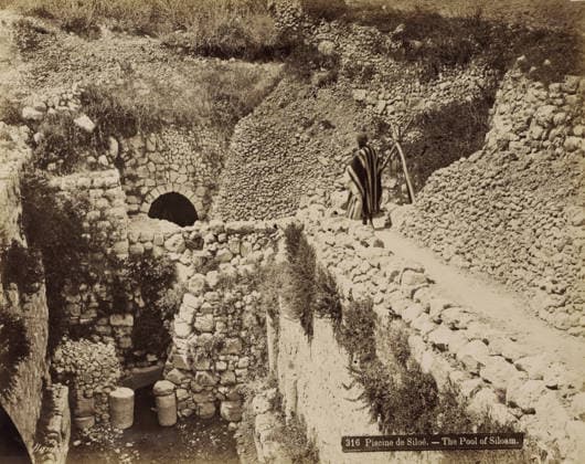 RECORD DATE NOT STATED Pool of Siloam near Jerusalem, Piscine de Siloà . - The Pool of Siloam., photograph, Part of Travel Album with Photos of Jerusalem and Other Biblical Places., negative number: 316 , photographer: Maison Bonfils, Jerusalem, c. 1867 - c. 1895, pasteboard, paper, albumen print, height 225 mm x width 283 mm, pond, pool, A sepia-toned scene of a rocky terrace and pathway carved into a hillside, showing rough stone walls and piled stones forming terraces and retaining structures, with an arched stone opening set into one wall above a lower chamber that contains large cylindrical stone columns partially submerged in water; vegetation and scrub grow among the stones and on the hillside, and a person wearing a striped cloak and head covering stands Copyright Penta Springs LCK260531_18255