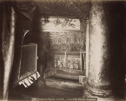 RECORD DATE NOT STATED Crib in the Cave of the Nativity in Bethlehem, Grotte de la Nativite: la creche.- Cave of the Nativity: the manger., photograph, Part of Travel Album with Photos of Jerusalem and Other Biblical Places., negative number: 893 , photographer: Maison Bonfils, Bethlehem (Israel), c. 1867 - c. 1895, Palestine, pasteboard, paper, albumen print, height 222 mm x width 282 mm, cradle, crib, A dim interior scene with patterned fabric draped across the ceiling and walls, a hanging ornate lamp near the top center, a painted panel showing figures and clouds partially visible above the fabric, a decorative tapestry with intricate motifs covering the back wall, a metal mesh enclosure with objects behind it in the middle foreground, heavy textured columns Copyright Penta Springs LCK260531_18260