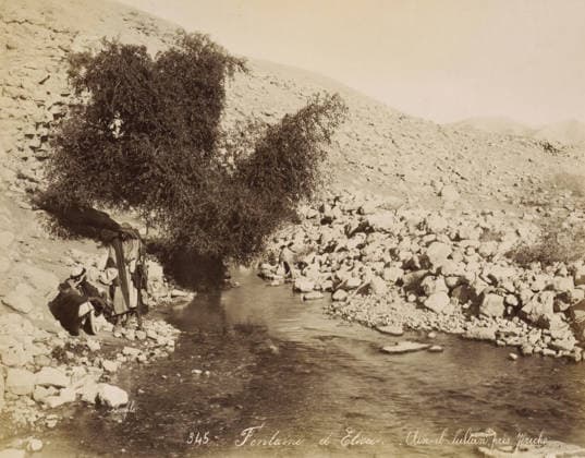 RECORD DATE NOT STATED Men at the well Ein as-Sultan near Jericho, Fontaine d Elisee. - Aïn-el-Sultan, pres Jà richo, photograph, Part of Travel album with photos of Jerusalem and other biblical places., negative number: 345 , photographer: Maison Bonfils, Jericho, c. 1867 - c. 1895, Maison Bonfils, photographer, Palestine, West Bank, pasteboard, paper, albumen print, height 223 mm x width 281 mm, well, A rocky riverside scene with a narrow stream flowing toward the foreground, smooth water reflecting light and rippling around scattered stones, a large leafy tree with dense foliage standing near the water s edge and casting a shadow over part of the stream, a hillside covered with loose rocks rising behind the tree and extending across the background, several Copyright Penta Springs LCK260531_18264
