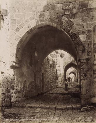 RECORD DATE NOT STATED JÃ rusalem - Ruelle allant au palais d HÃ rode, Street to Herod s Palace in Jerusalem, photograph, Part of Travel album with photos of Jerusalem and other biblical places., negative number: 259 , photographer: Maison Bonfils, Jerusalem, c. 1867 - c. 1895, pasteboard, paper, albumen print, height 282 mm x width 222 mm, arch, archivolt, architecture, sloping street, street with steps or stairs, A narrow cobblestone passageway lined with rough stone walls and successive stone arches receding into the distance is shown, with light filtering through the arch openings and a hanging lantern on the left wall under the nearest arch; a person wearing a long garment stands facing away in the middle distance while another figure is farther along under Copyright Penta Springs LCK260531_18269