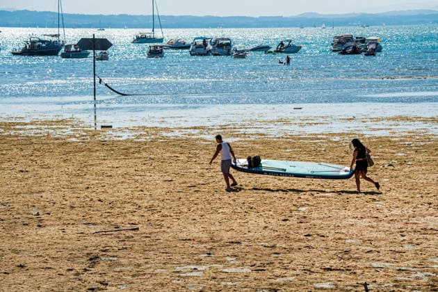 Zwei Wassersportler tragen ihr großes Stand Up Paddle Board durch die breite trockengefallene Uferzone zum Wasser, Wasserburg, August 2026 Deutschland, Wasserburg, August 2026, Niedrigwasser am Bodensee, zwei Wassersportler tragen ihr großes Stand Up Paddle Board durch die breite trockengefallene Uferzone zum Wasser, durch Trockenheit freigelegte Uferzone am Bodensee,, die aktuelle Trockenheit hat den Pegelstand stark sinken lassen, die Boote liegen weit draußen, die Schifffahrt ist eingeschränkt, Niedrigwasser, Wasserstand sehr niedrig, zu wenig Regen, schönes Wetter, Hochsommer, Hitzewelle mit Temperaturen bis 33 Grad, Sommer, Landkreis Lindau, Bayern,