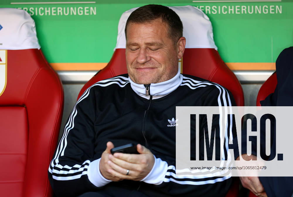 Fussball, Herren, Saison 2025 2026, FC Augsburg - FC Bayern München, Bundesliga, WWK Arena Max Eberl, Sportvorstand (FC Bayern München) sitzend auf der Trainerbank mit Blick auf das Handy, Smartphone, Telefon, grinsen, schmunzelnd DFL DFB regulations prohibit any use of photographs as image sequences and or quasi-video