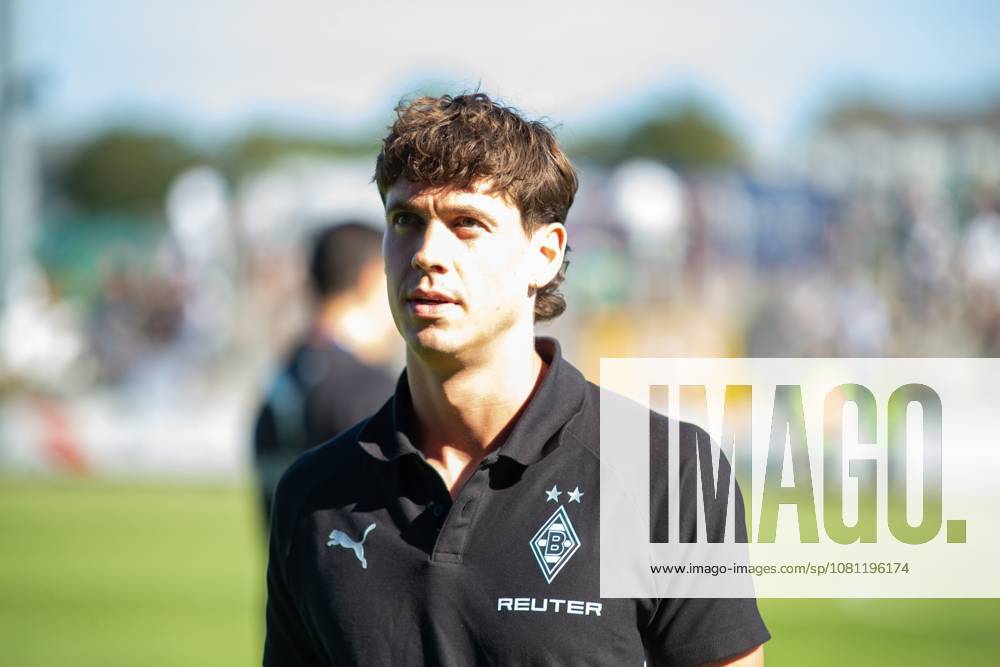 Ein Offizieller von Borussia Mönchengladbach steht im schwarzen Vereins-Polo mit Puma-Logo und REUTER-Sponsoring vor dem Spielfeld. Im Hintergrund sind Zuschauer und das Spielfeld zu erkennen. Borussia Mönchengladbach – Offizieller in Velbert