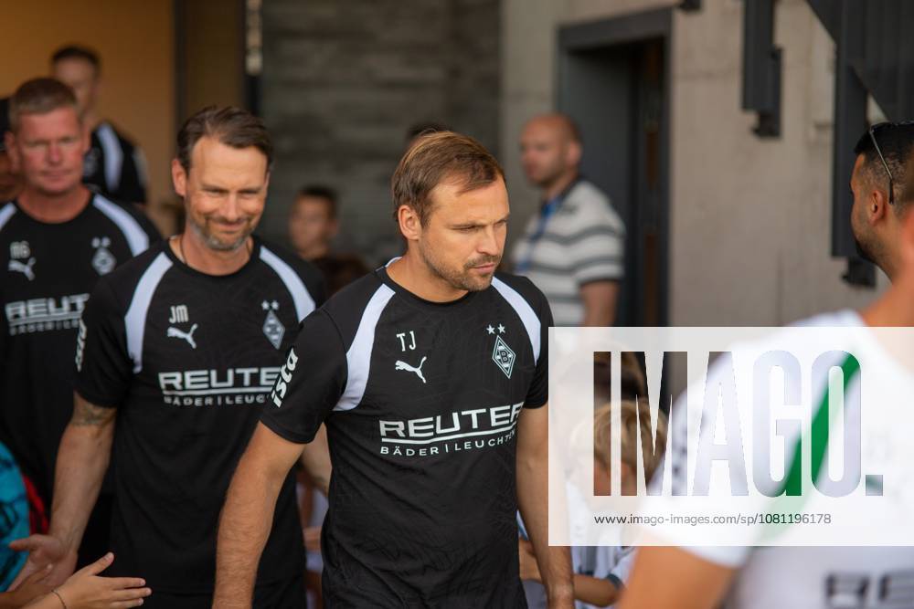 Zwei Mitglieder des Trainerteams von Borussia Mönchengladbach in schwarzen Trainingsshirts mit REUTER-Sponsoring laufen durch einen Eingang, umgeben von Fans und Kindern. Die Initialen TJ und JM sind auf den Shirts sichtbar. Borussia Mönchengladbach Trainerteam beim Einlaufen in Velbert