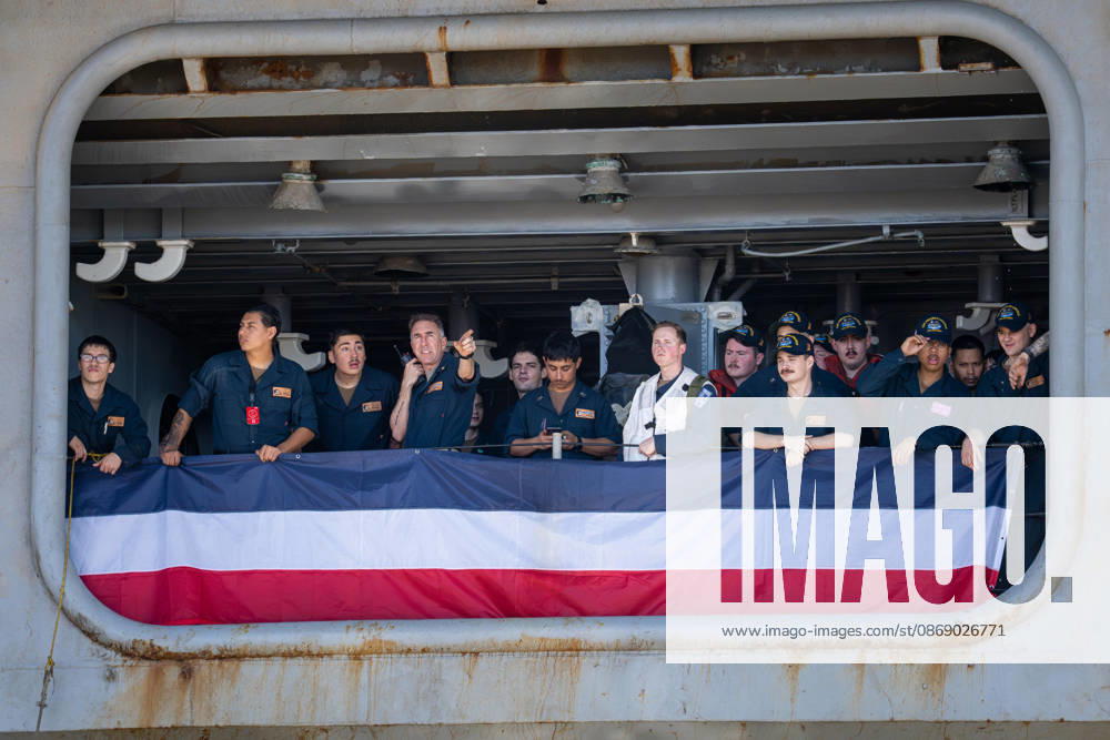 U.S. Navy sailors stand behind red, white and blue bunting aboard the aircraft carrier USS Abraham Lincoln (CVN 72) as the ship returns to Naval Air Station North Island in Coronado, California, on October 8, 2026, following a deployment. One sailor points toward the shore as others look on. Coronado United States - ZUMAg374 20261008_znp_g374_010